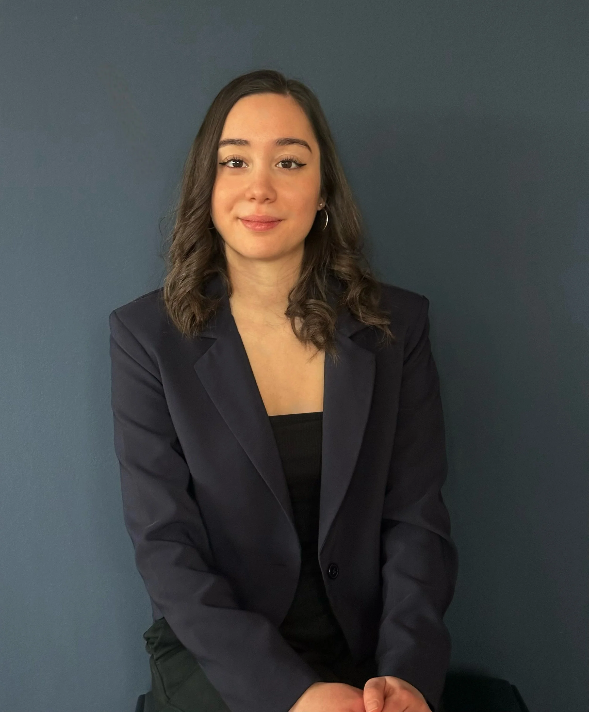 Junge Frau mit braunen, lockigen Haaren, dunklem Blazer und schwarzen Oberteil vor einer blauen Wand.