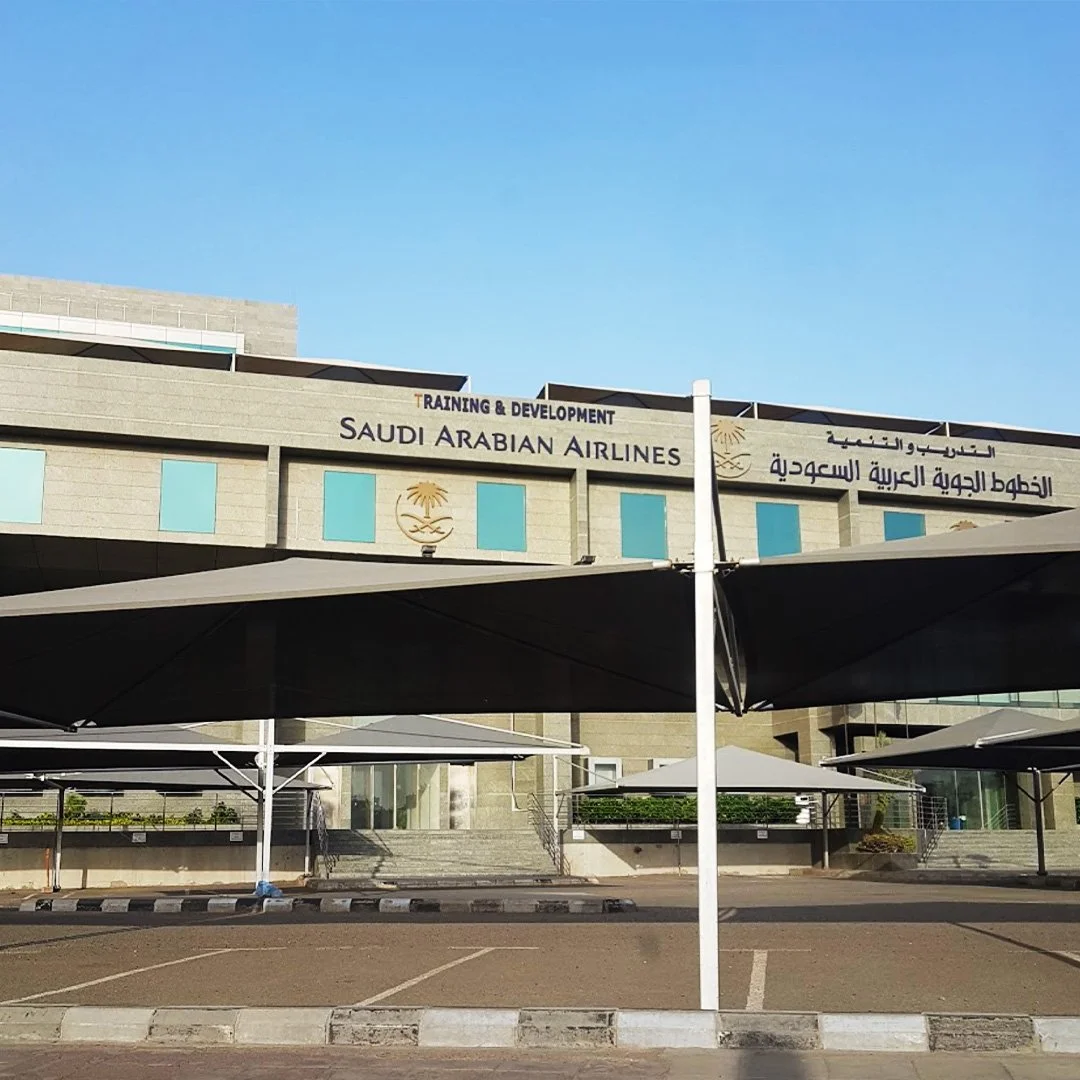 Exterior view of a building with signs for Saudia Airlines and Training & Development, with shaded parking areas in front. مبنى الخطوط السعودية