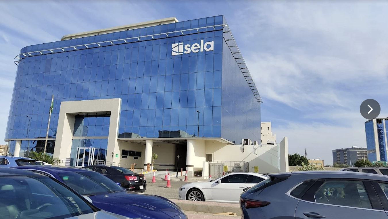 A modern glass office building with the logo 'Sela' on the top right corner, surrounded by a parking lot with several cars parked. مبنى تجاري في طريق الملك في جدة