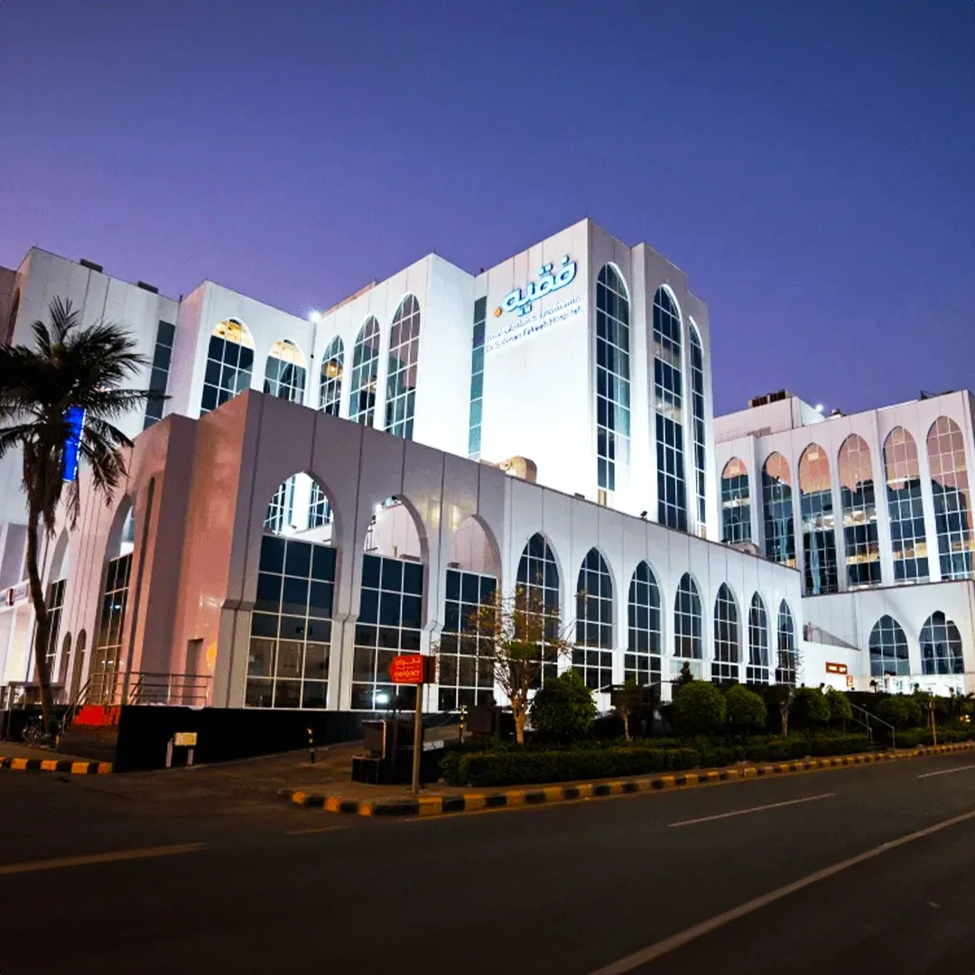 A large white multi-story building with tall arched windows illuminated at night, with trees and street in foreground. مستشفى فقيه في جدة