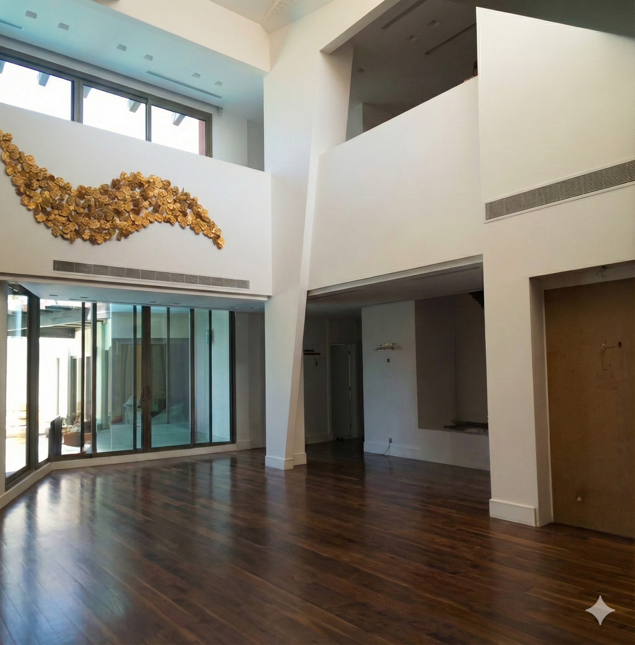 Interior of a modern, spacious living area with hardwood floors, white walls, large windows, and an abstract gold wall sculpture. Interior design of a beautiful villa in Jeddah. تصميم داخلي فيلا في جدة