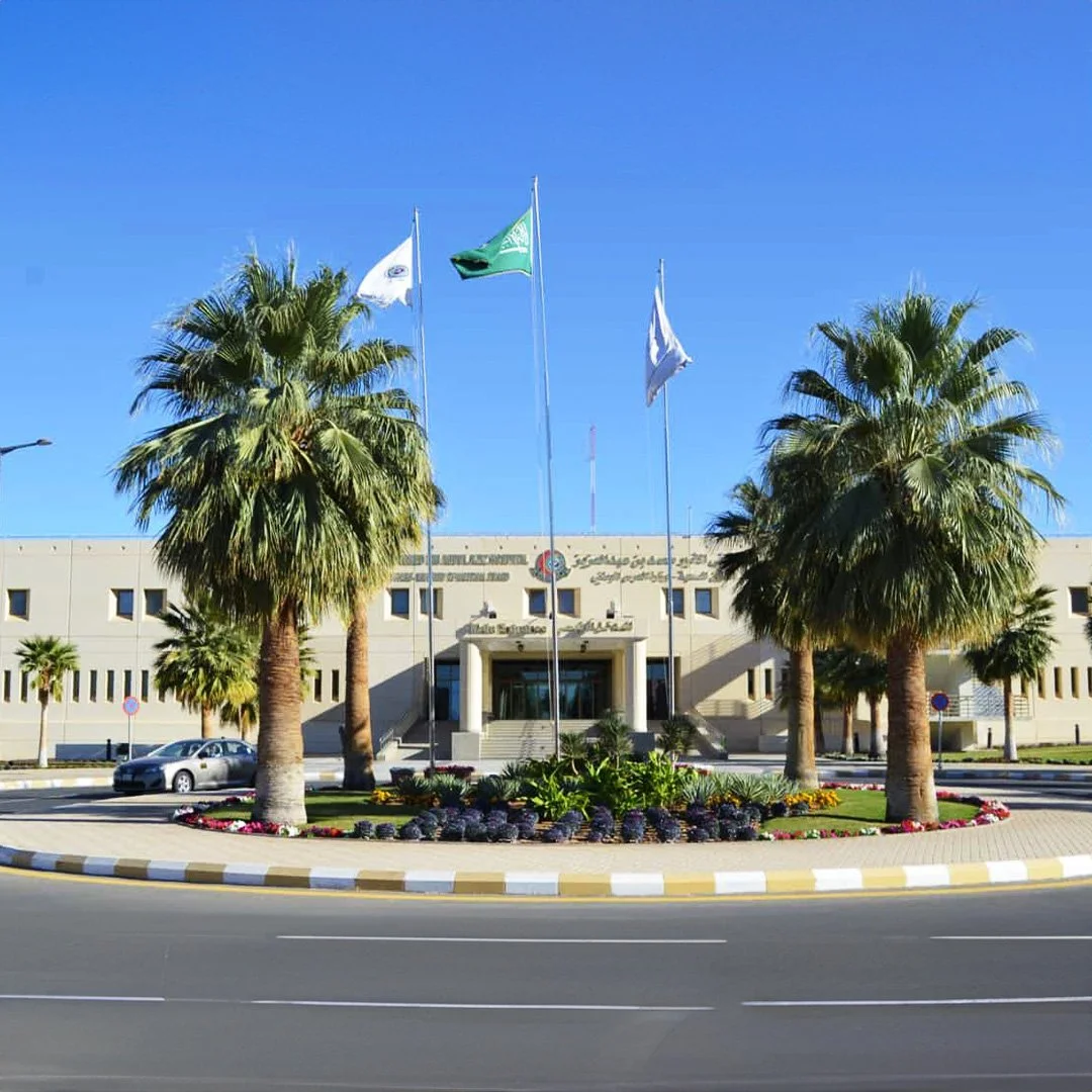 King Abdulaziz Hospital building with Arabic and English signs, flanked by palm trees, with three flags on poles in front, including a green flag and two white flags, under a blue sky. مدخل مستشفى الملك عبدالعزيز