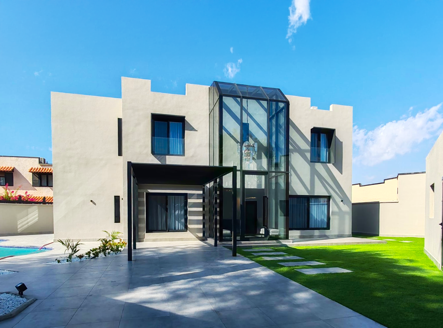 Modern two-story house with flat roof, large windows, and a glass-enclosed entrance, surrounded by a green lawn and a patio with a swimming pool, under a clear blue sky. بيت في أبحر في جدة