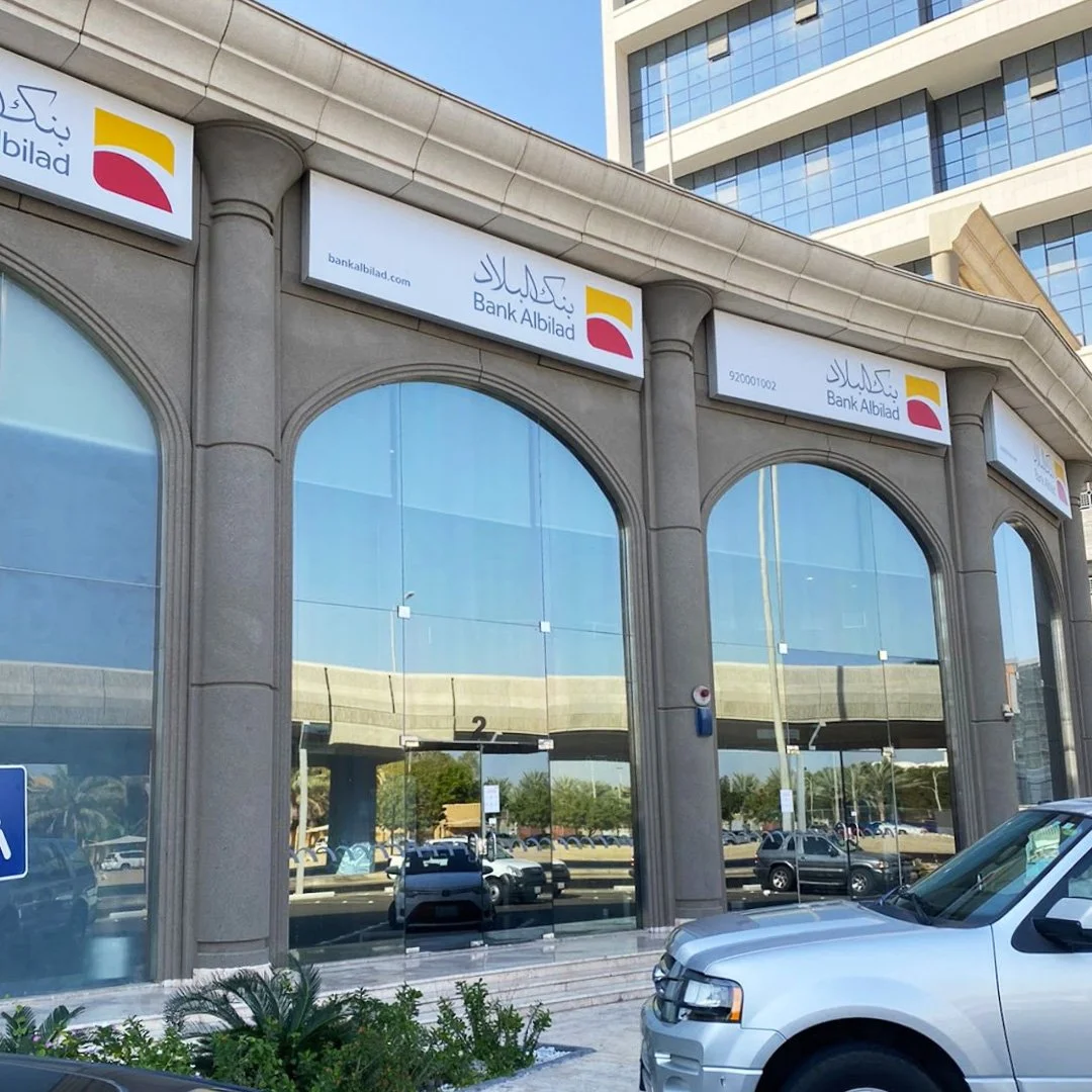 Facade of a bank building with glass windows, signage for Bank Albilad in Arabic and English, and parked cars in front. مبنى بنك البلاد في جدة حي الزهراء