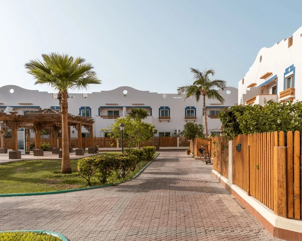 A sunny courtyard with palm trees, greenery, a paved walkway, wooden fencing, and white buildings with blue accents and balconies, resembling a Mediterranean or coastal resort. فلل البساتين في جدة