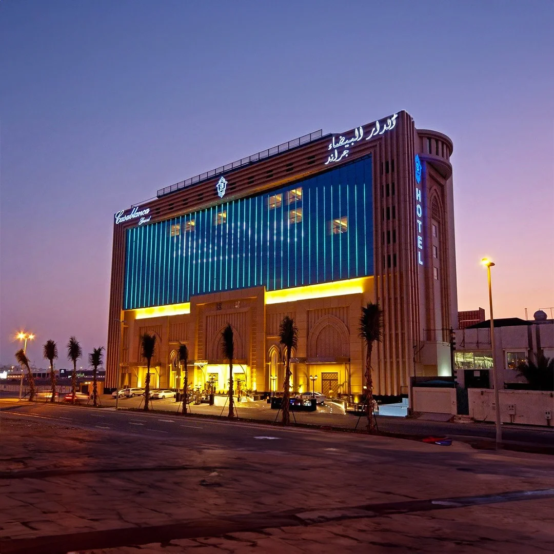 Large hotel building at dusk with blue and yellow lighting, palm trees in front, and a clear purple sky. فندق الدار البيضاء في جدة. Casablanca Hotel in jeddah