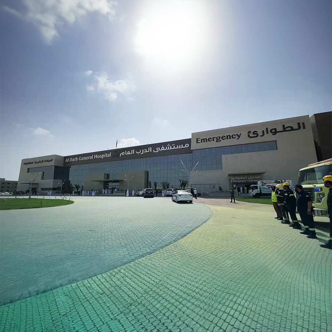 Front view of Al Darb General Hospital with emergency entrance, vehicles parked outside, and people standing nearby on a sunny day. مستشفى الدرب في جيزان