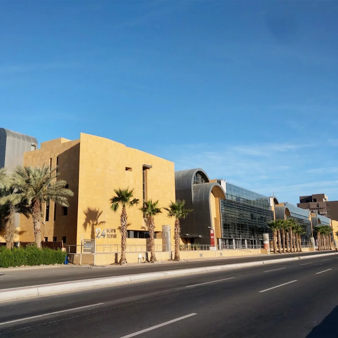 Modern building with a yellow facade, glass windows, and arched roofs, palm trees lining the sidewalk, and a clear blue sky in the background. سعودي ديار المقر الرئيسي