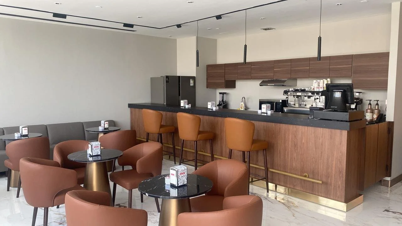 Modern cafe or lounge area with brown leather armchairs, small black marble-topped tables, and a wooden bar with barstools. The bar has a black countertop, with kitchen appliances and coffee supplies behind it. The room has a light-colored marble floor and minimalistic decor.