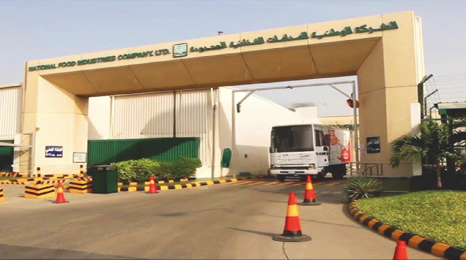 Entrance to the National Food Security Company Ltd. with a white truck parked inside, orange and black traffic cones, and greenery surrounding the area. Luna. الشركة الوطنية لصناعة الغذائيات في السعودية