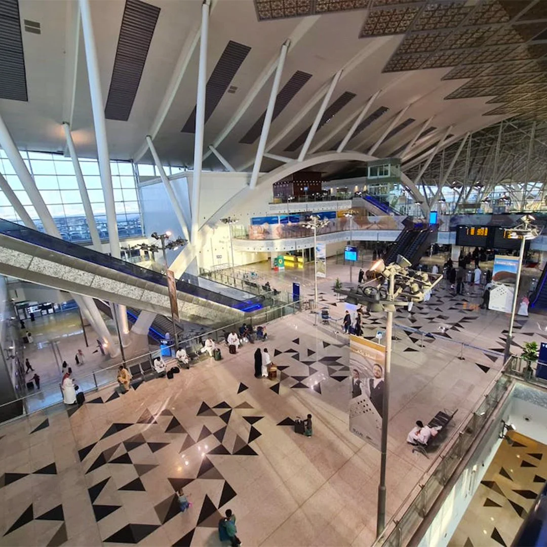 Interior of Mekkah train station with high ceiling, large windows, escalators, and seating areas. Passengers are sitting and walking around. محطة قطار مكة
