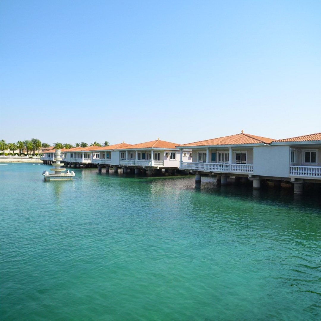 Waterfront row of houses with orange-tiled roofs on stilts over water, palm trees in background, clear blue sky. فلل أبحر