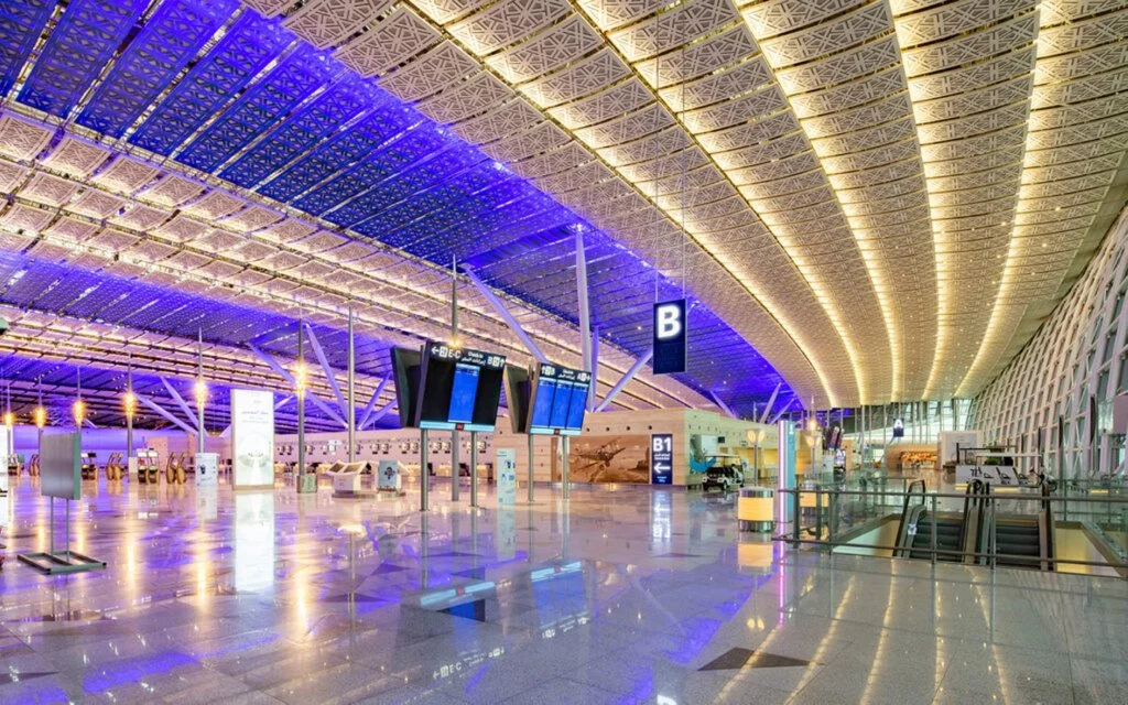 Empty airport terminal with colorful lighting, digital flight information displays, check-in counters, and escalators. مطار الملك عبد العزيز الدولي في جدة. Jeddah International Airport.