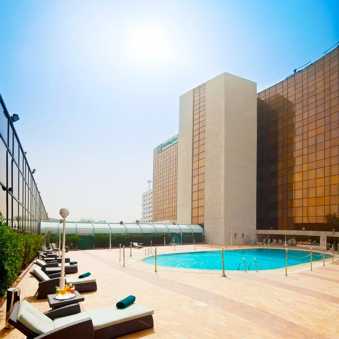 Hotel rooftop pool area with lounge chairs, small tables, and a swimming pool, surrounded by glass barriers and high-rise buildings under a clear blue sky. هوليداي ان طريق المدينة