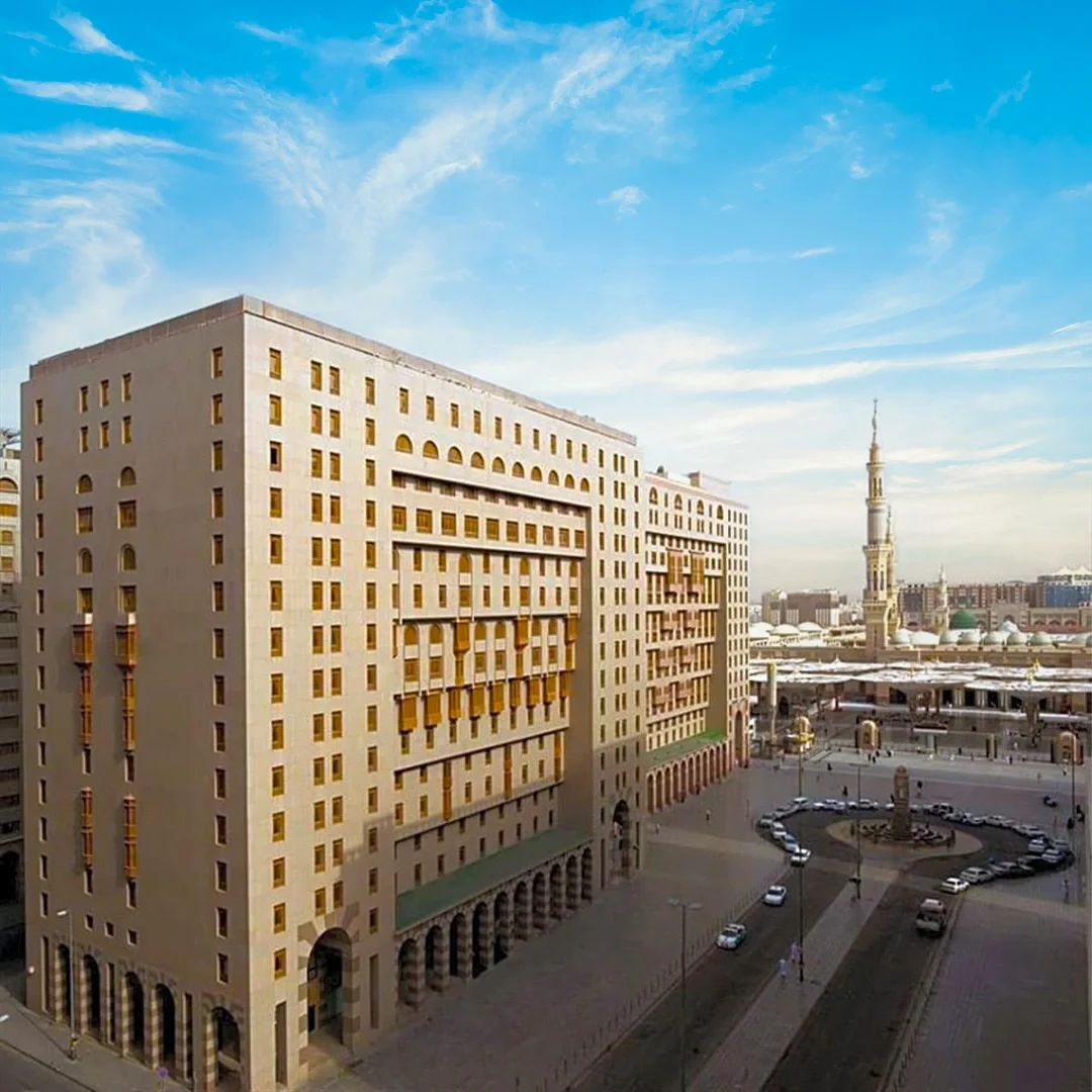 A large beige building with multiple small windows and archways on the ground level, in an urban setting with a circular driveway, parked cars, and a clock tower in the background under a blue sky with wispy clouds. Shaza AlMadinah Hotel in AlMadinah