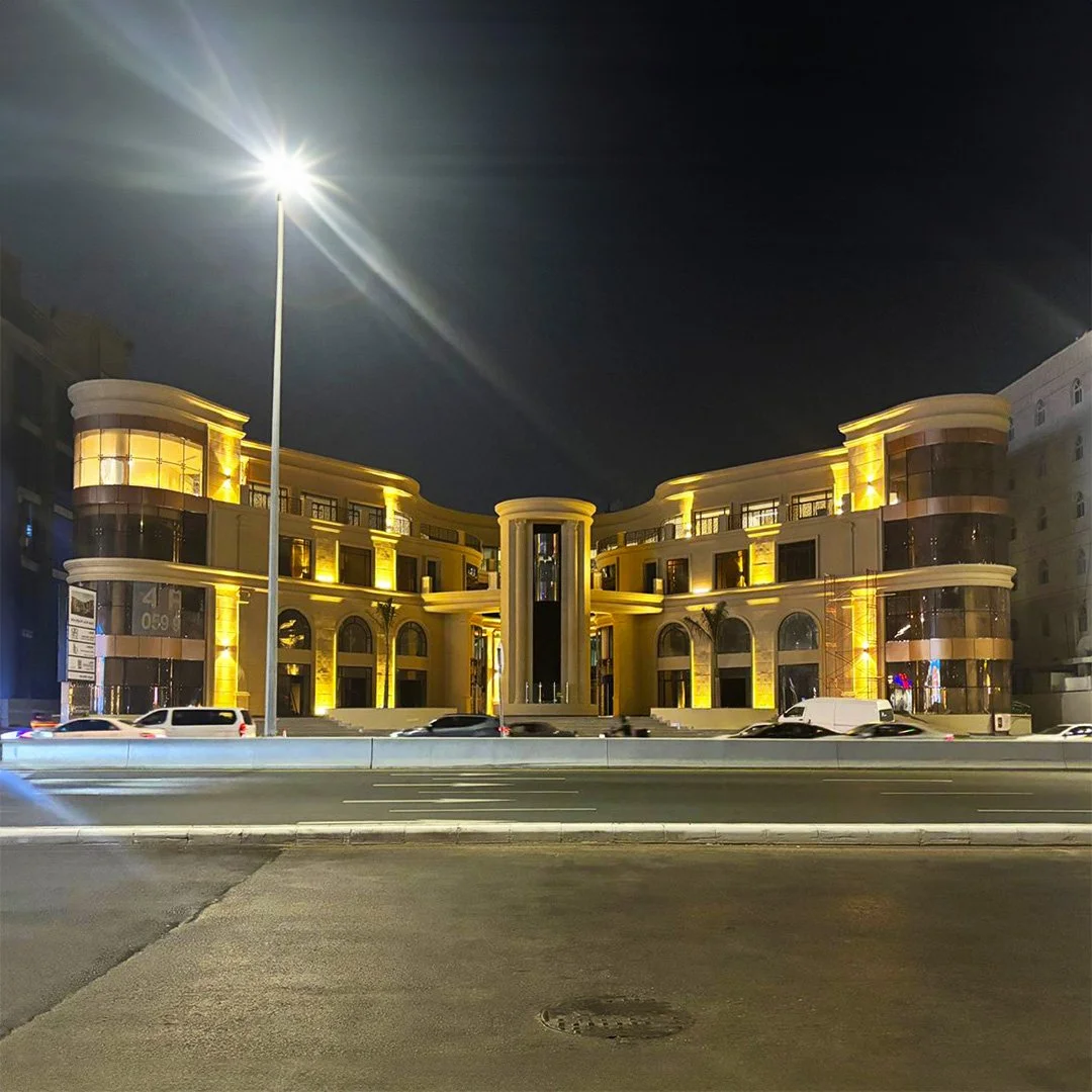 Night view of a modern, well-lit building with rounded edges and glass balconies, illuminated with warm yellow lights, situated along a multi-lane road with cars passing by, under a tall streetlight, Al Tahlia Road in Jeddah Saudi Arabia