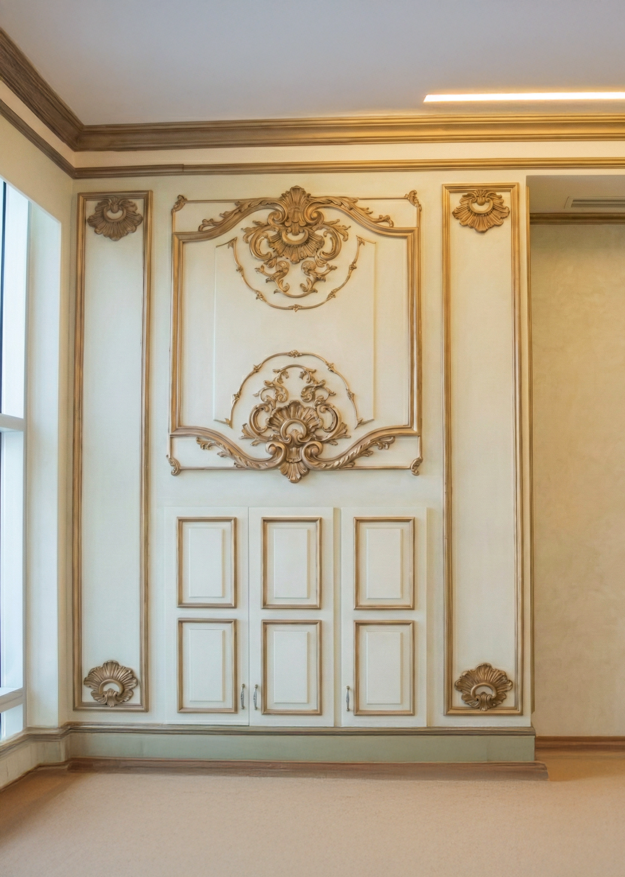 Elegant interior wall of a studio with ornate gold decorative moldings and a built-in cabinet with six panels, enhanced by crown molding and a neighboring window on the left. ستوديو
