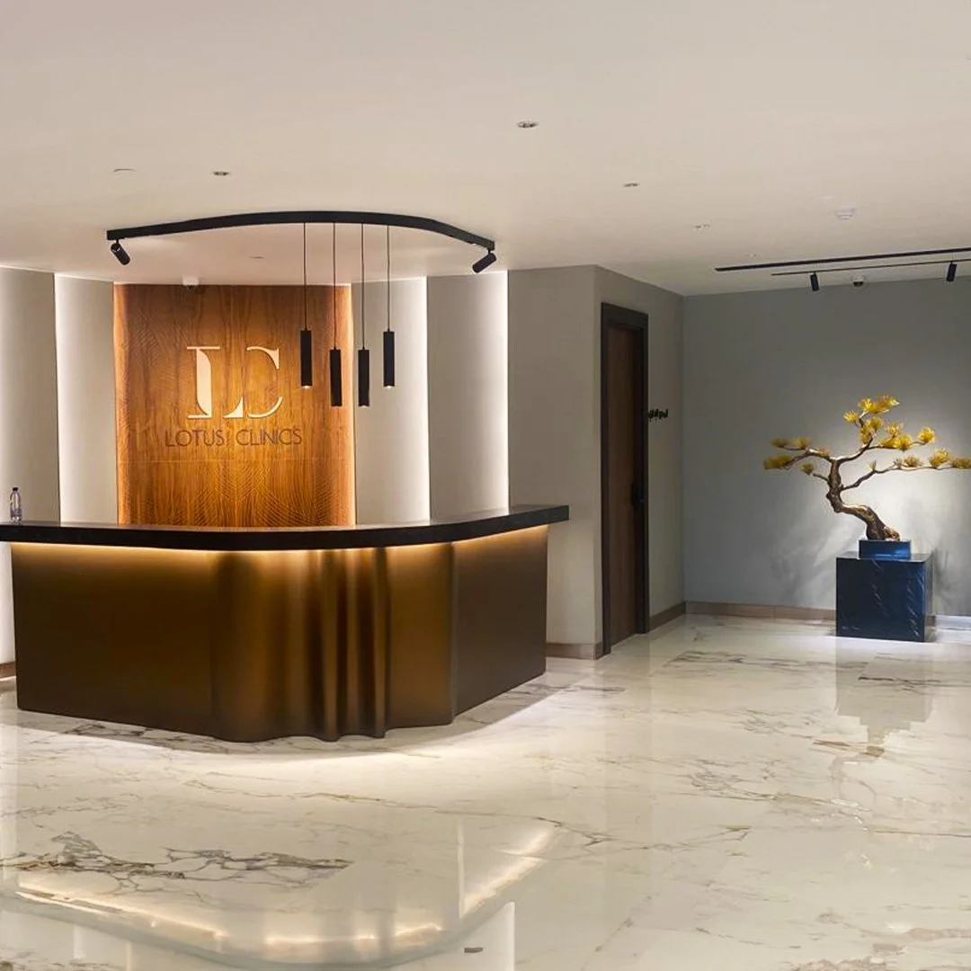 Modern reception area with marble floor, lotus clinics sign on wooden wall, black and gold curved counter, black hanging lights, and a decorative bonsai tree in a black planter. عيادات لوتس الطبية في جدة