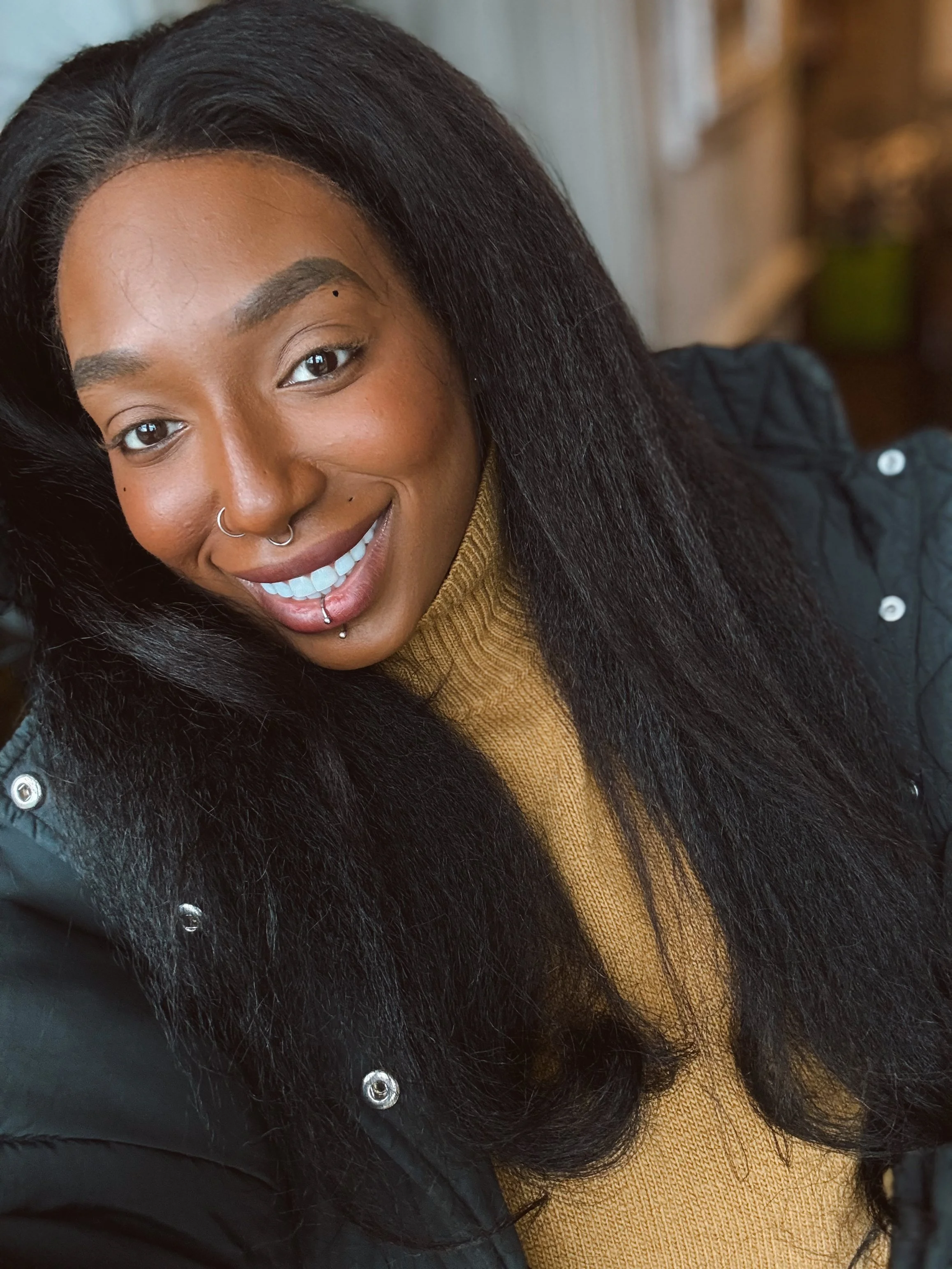 A young woman with dark skin, long black hair, and a nose and lip piercing, smiling at the camera. She is wearing a mustard yellow turtleneck and a black jacket with silver buttons.