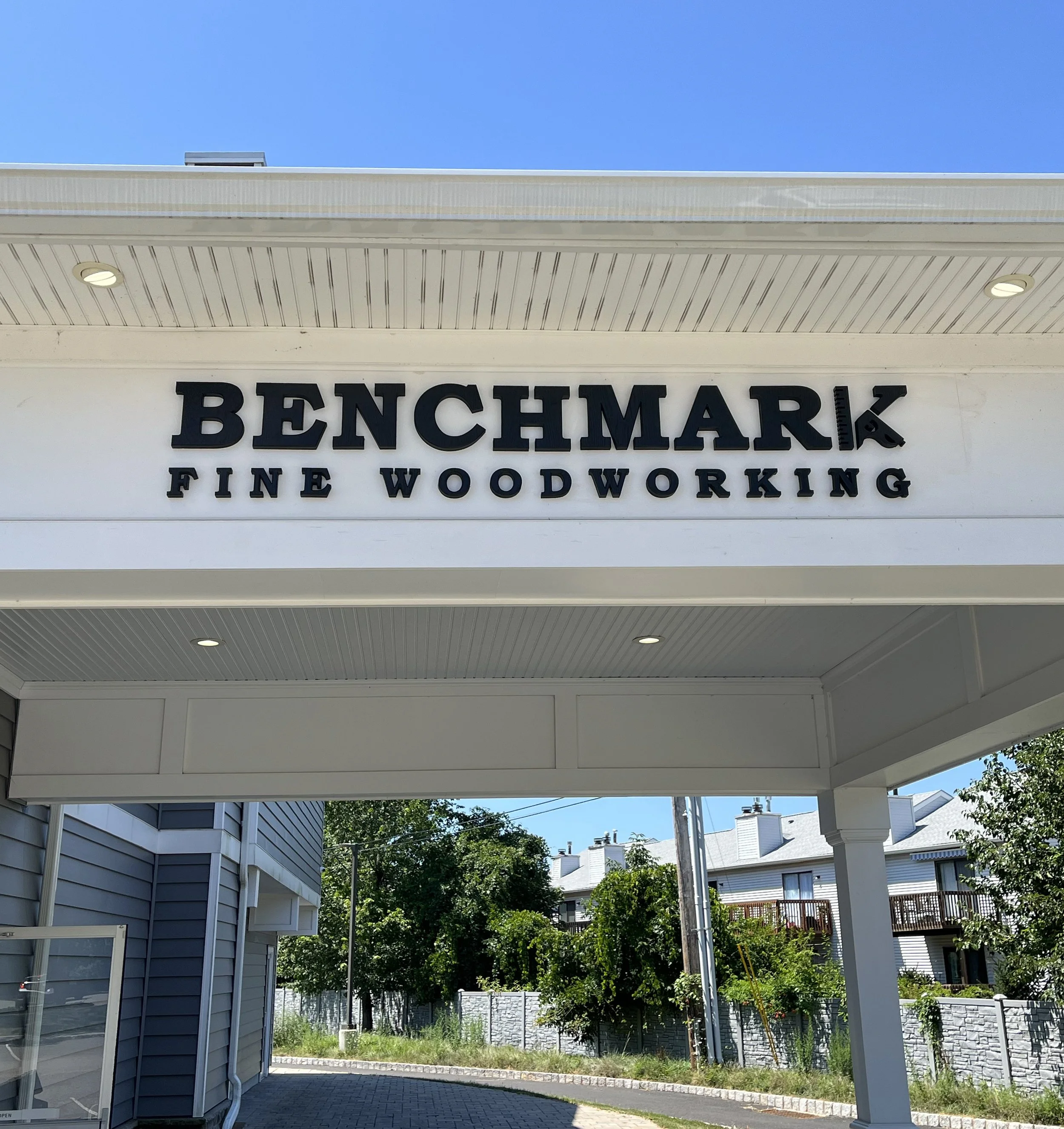 Sign for Benchmark Fine Woodworking store with black letters on a white background, situated outdoors in a suburban area with residential buildings and trees in the background.