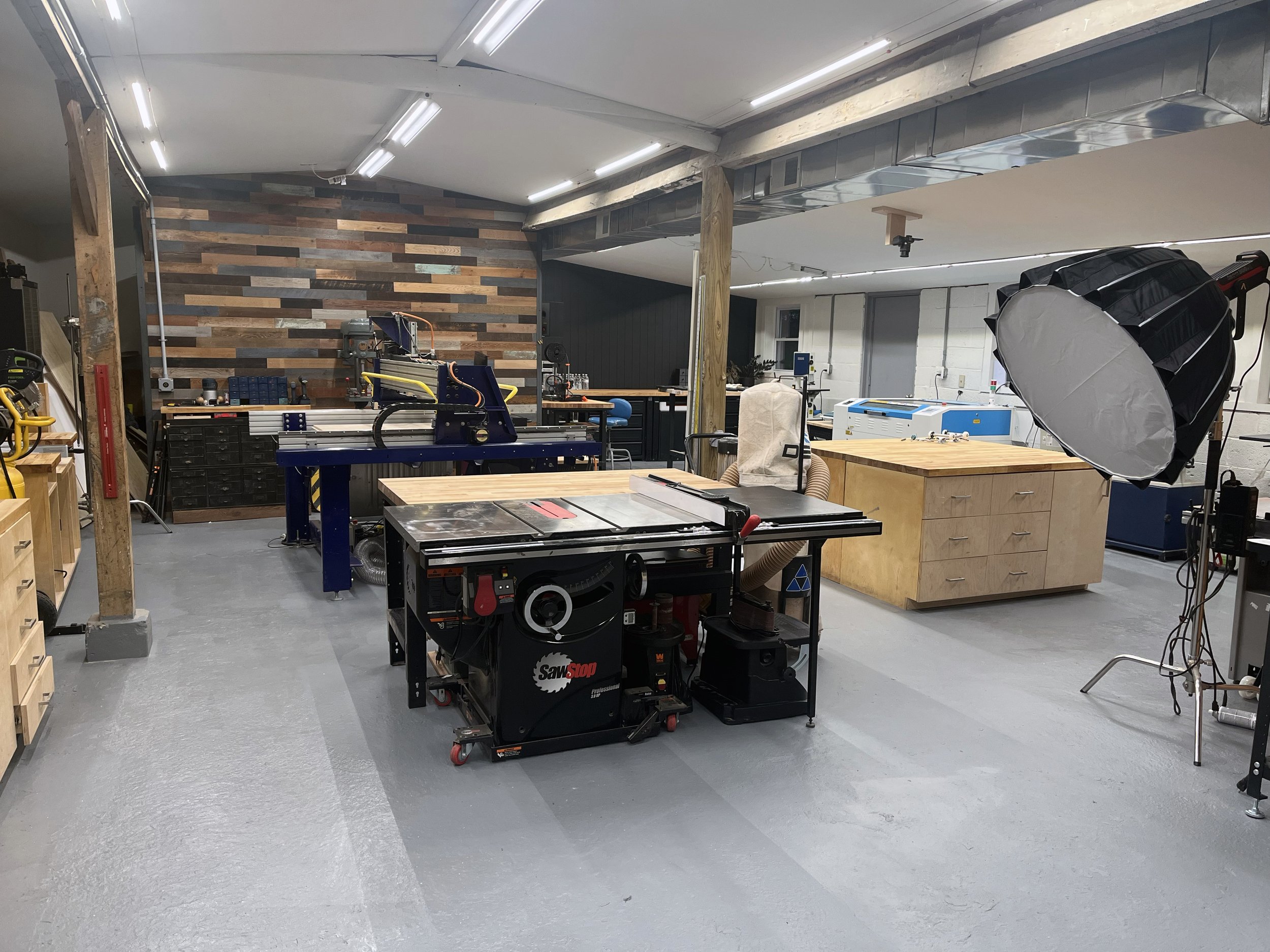 A workshop with woodworking tools, a SawStop table saw, workbenches, storage drawers, a large photography softbox, and equipment, with a wooden accent wall and industrial lighting.
