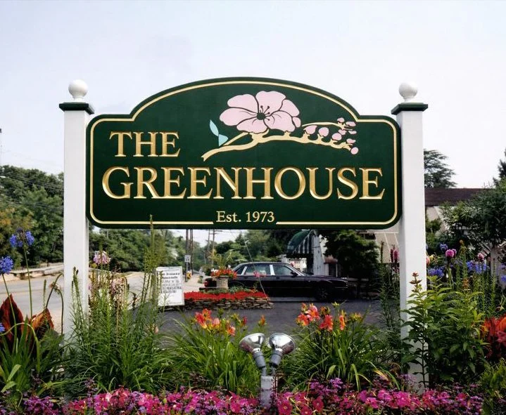 Sign for The Greenhouse with a flower illustration, established in 1973, surrounded by colorful flowers and plants.