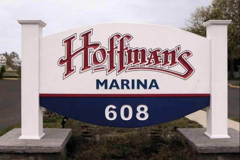 Sign for Hoffman’s Marina with the address 608, featuring a white background, red and blue text, and mounted on a white post with stone bases.
