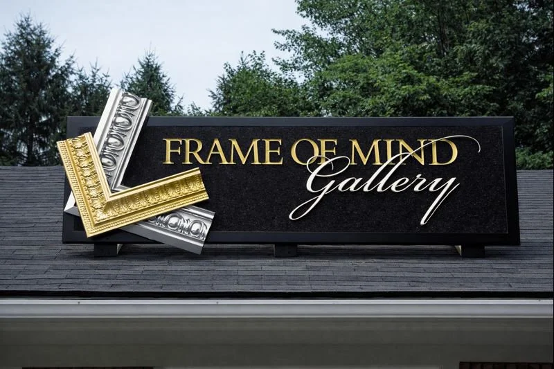 Sign reading "Frame of Mind Gallery" with decorative gold and silver frame accents, located on the roof of a building with trees in the background.