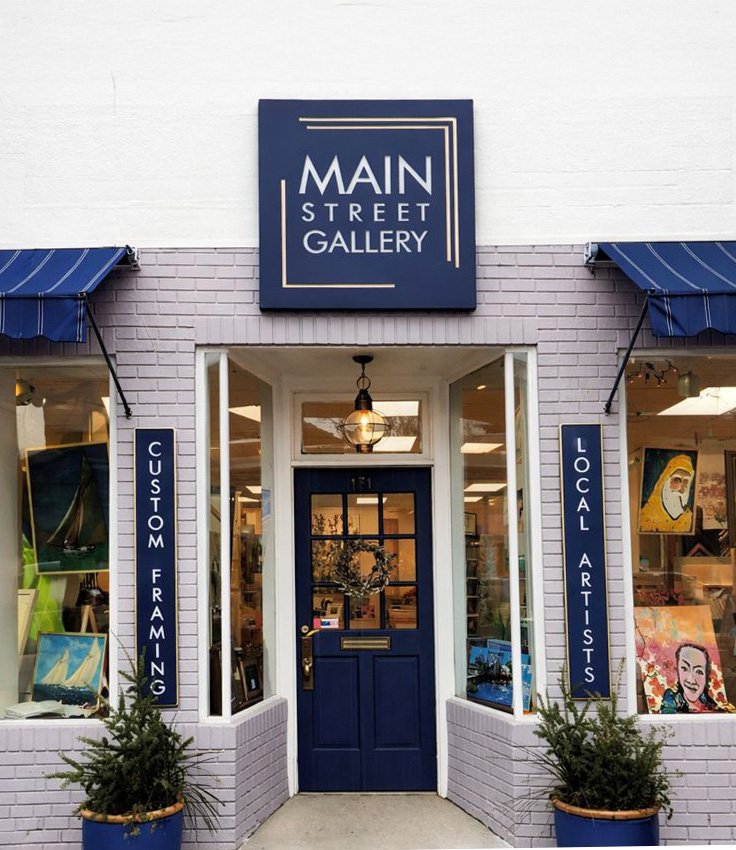 Storefront of Main Street Gallery featuring a blue door, large windows on either side displaying artwork, and signage indicating custom framing and local artists.
