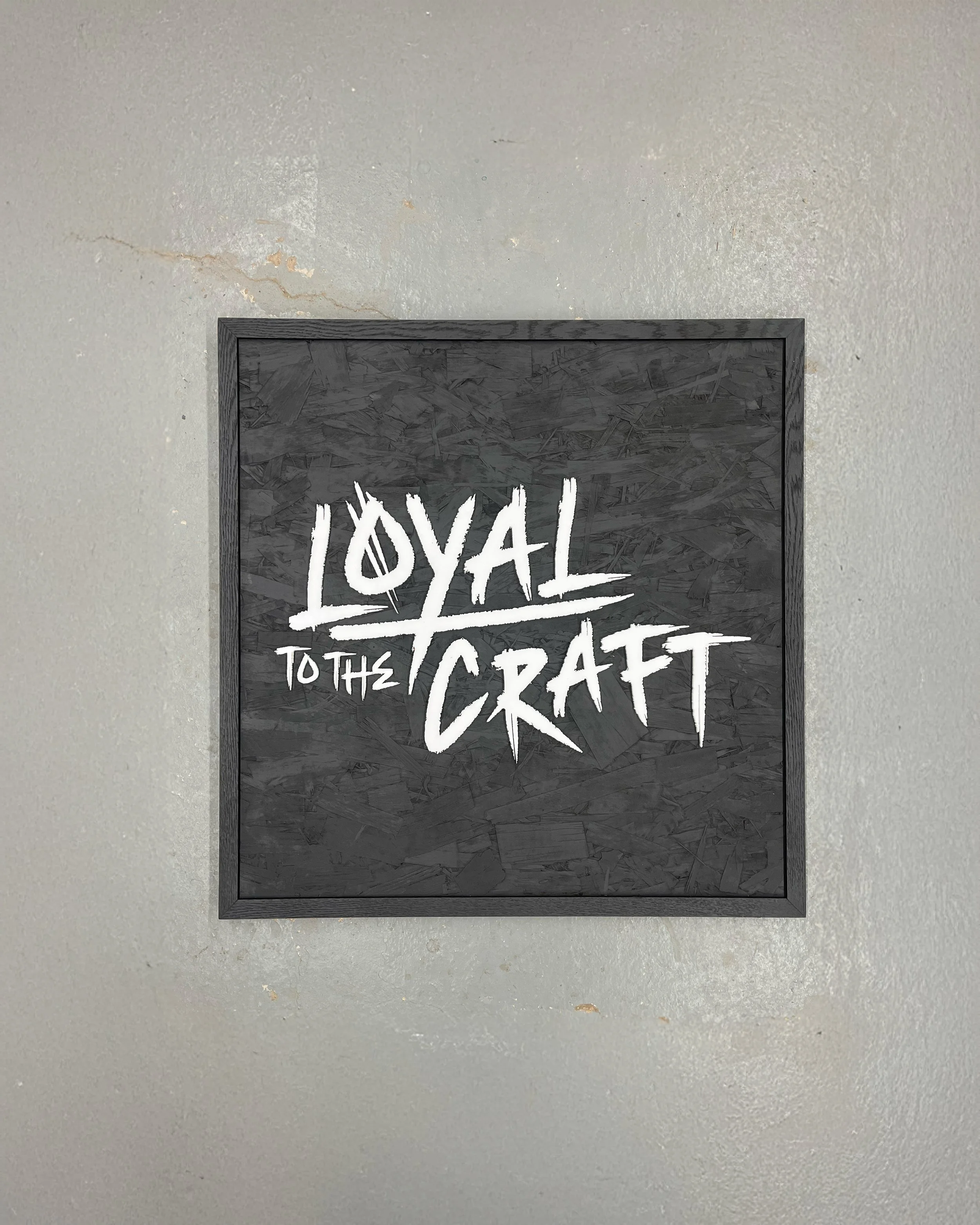 A square black sign with white handwritten-style text that says 'Loyal to the Craft,' mounted on a gray wall.