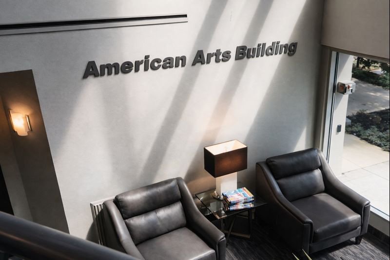 Interior view of the American Arts Building with two black leather armchairs, a side table with a lamp and magazines, a wall sign reading "American Arts Building," and a large window letting in natural light.