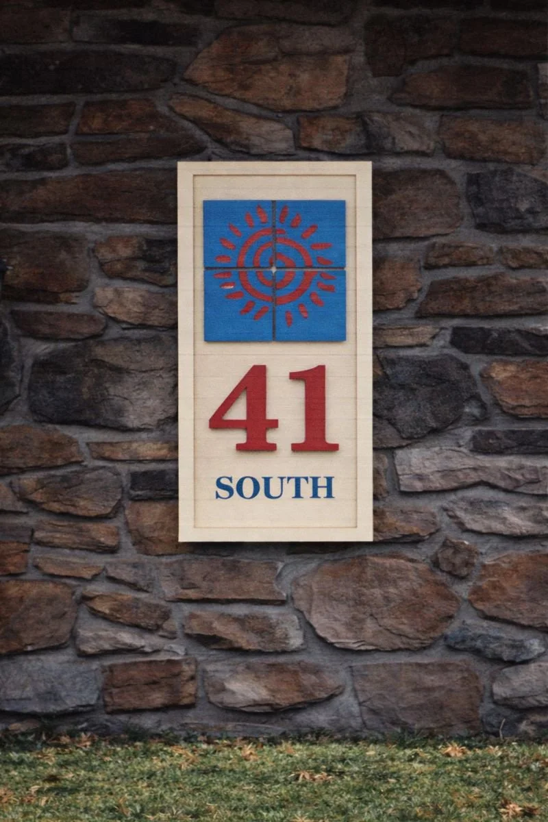 A house number sign on a stone wall with the number 41 in red, a blue square with a red spiral and sun design above the number, and the word SOUTH in blue below the number.
