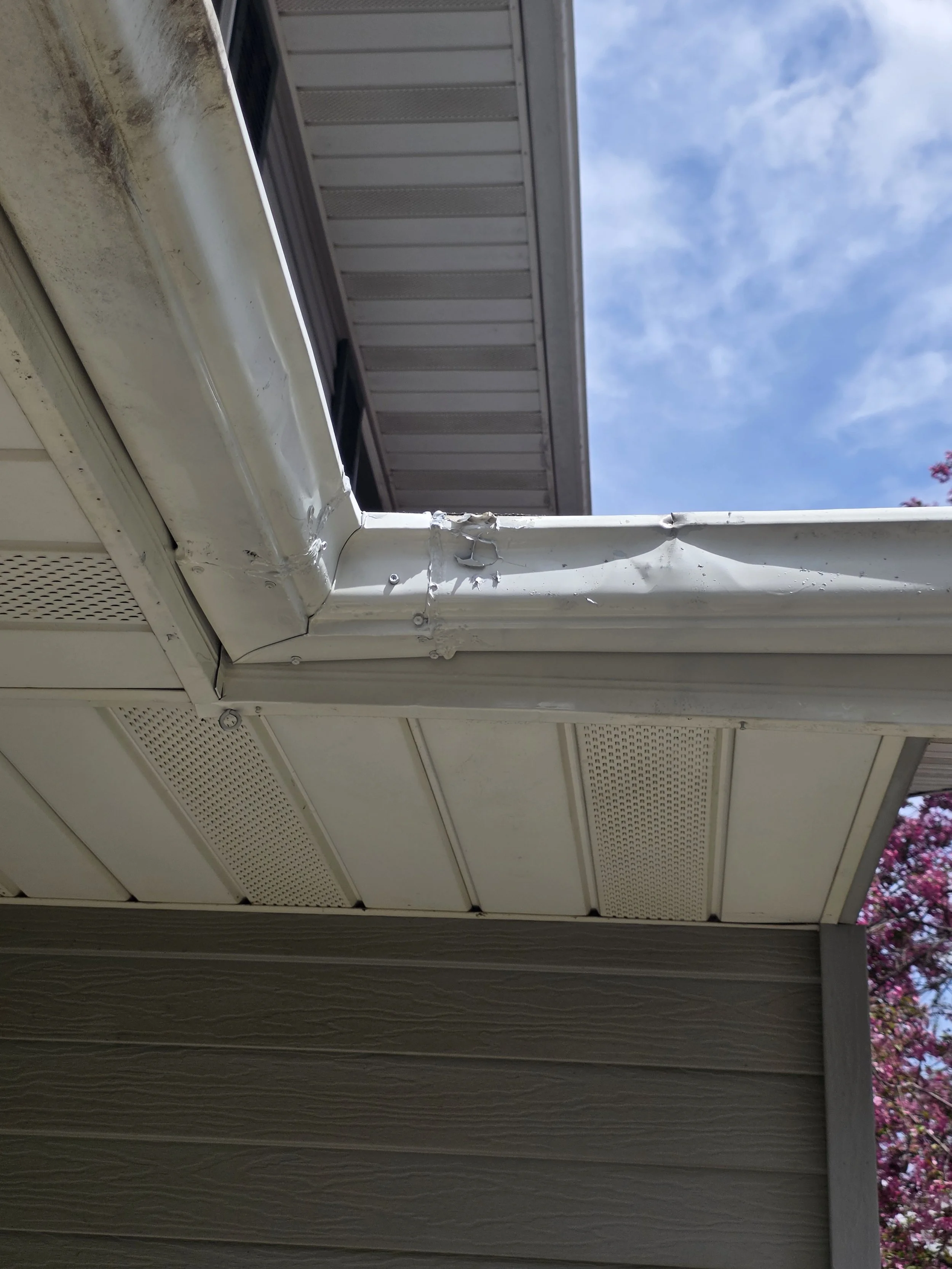 Close-up of a house's gutter and fascia board with some damage, seen from below and to the side. The sky with clouds is visible in the background.