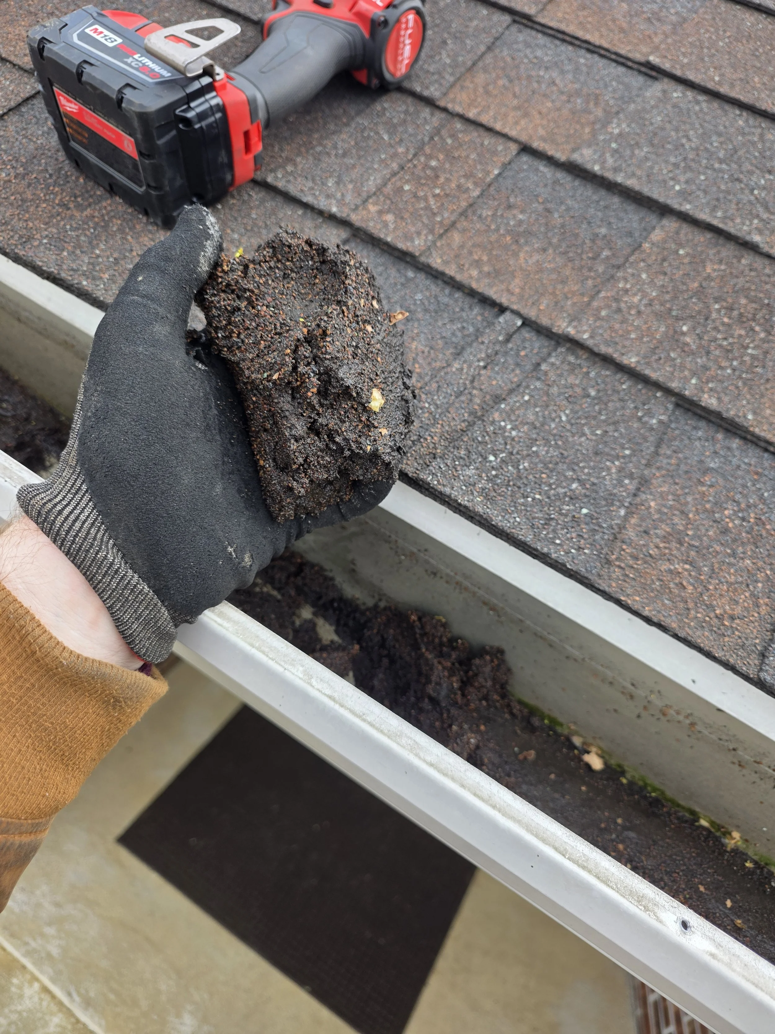 A hand wearing a black work glove holding a handful of soil or dirt. In the background, a cordless power drill is lying on a shingled roof, possibly for roofing work.