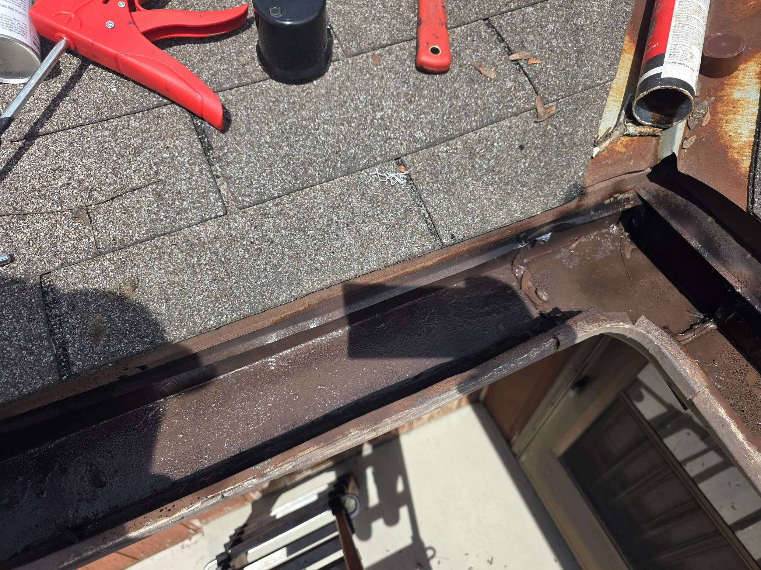 Tools and materials on a concrete patio, including a red coping clamp, a red handle, a black container, a foam can, and a rusty metal pipe, with part of a brick wall and a metal railing visible.