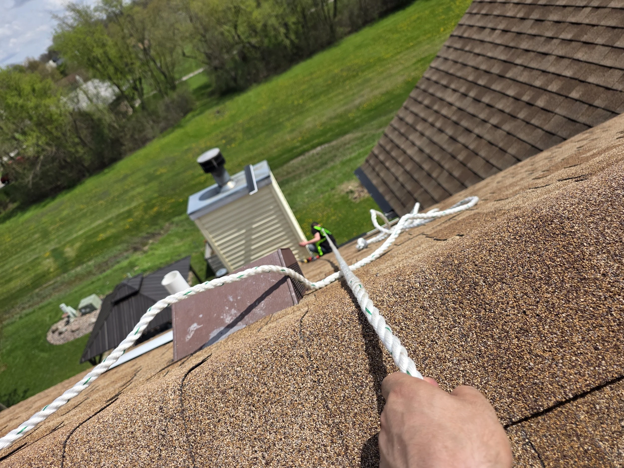 View from a roof showing a white rope, a chimney, and a person's hand in the foreground, with a green yard and trees in the background.