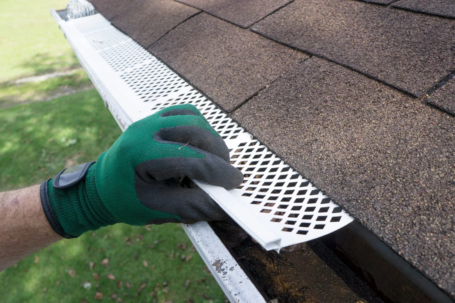 A person's hand wearing a green and black glove is installing white attic vent covers along the edge of a house roof with brown shingles.