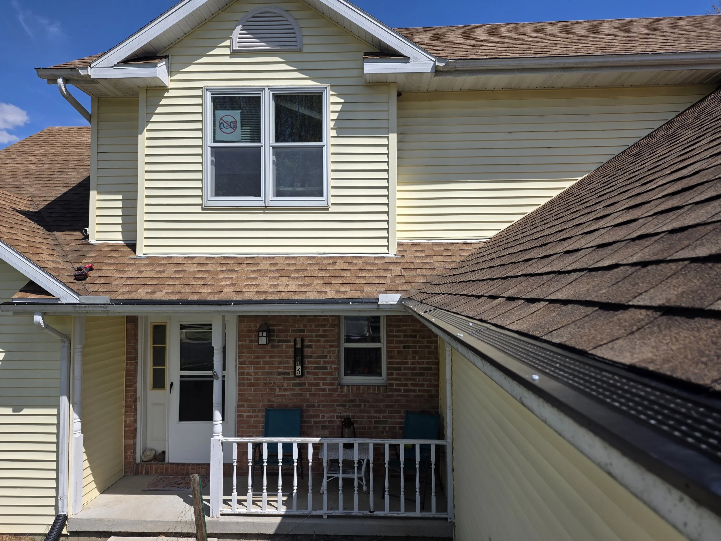The back porch of a yellow house with a brown shingle roof, white trim, a white front door, and a small patio area with outdoor chairs.