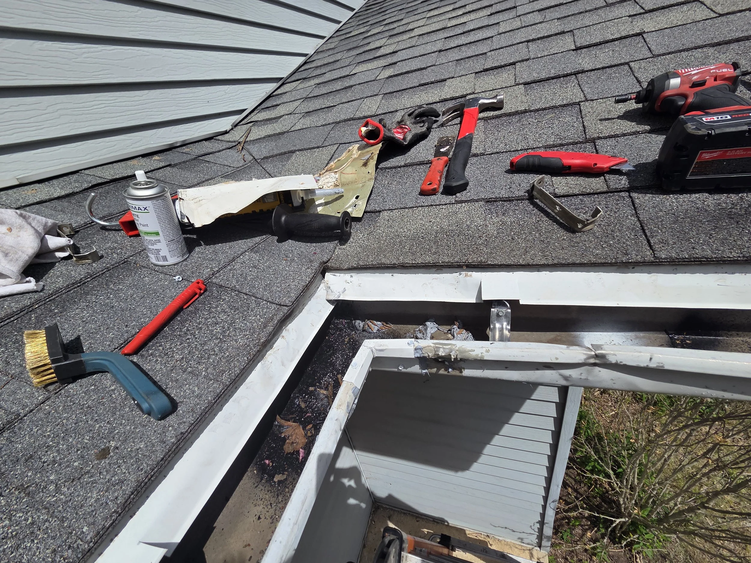 Tools and materials spread out on a rooftop, including a caulking gun, a cordless drill, a hand saw, a tape measure, a can of paint, and other construction tools, indicating roof repair or installation work.