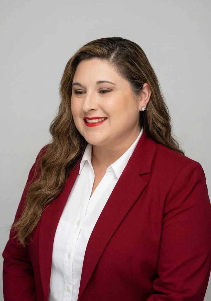 Portrait of a couple counseling specialist with long wavy hair wearing a red blazer, white shirt, and diamond earrings, smiling against a plain light gray background.