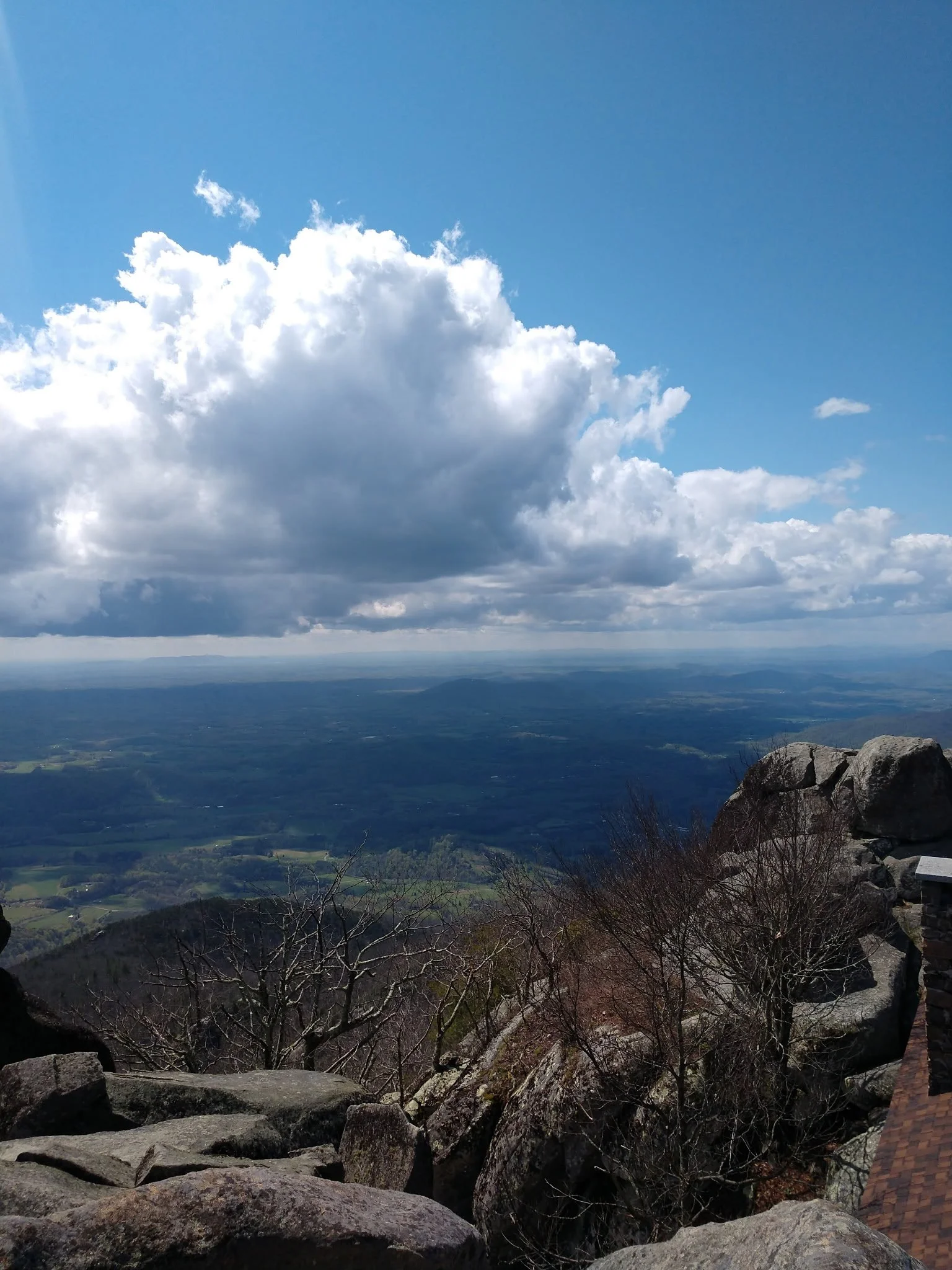 View from a mountain peak showing large rocks and leafless trees in the foreground, extending out to a broad landscape with green fields, and a sky with large, fluffy clouds.