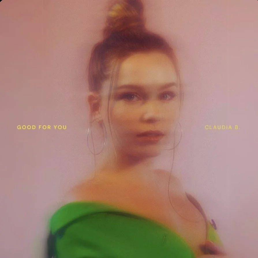 A woman with light skin and brown hair styled in a top bun with a yellow hair accessory, wearing large hoop earrings, a green top, and earbuds, standing behind a semi-opaque pinkish screen or glass with a soft-focused effect. Claudia B.