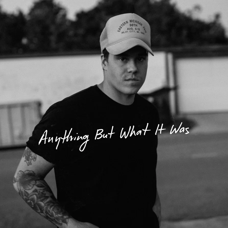 Black and white photo of a young man wearing a baseball cap and a black t-shirt, looking serious, outdoors during evening or late afternoon, with text overlay saying "Anything But What It Was." Zach John King