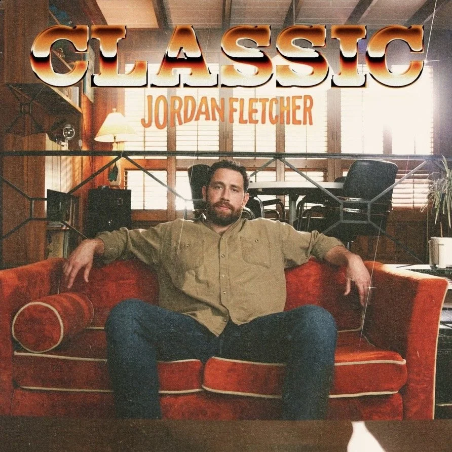 A man sitting on a red couch in a cozy wood-paneled room with large windows and blinds, with furniture and a lamp in the background. Jordan Fletcher