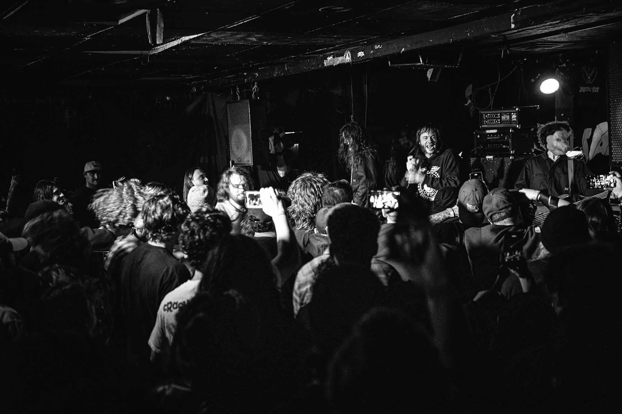 A black-and-white photo of a crowded concert with people on stage and an audience taking pictures.
