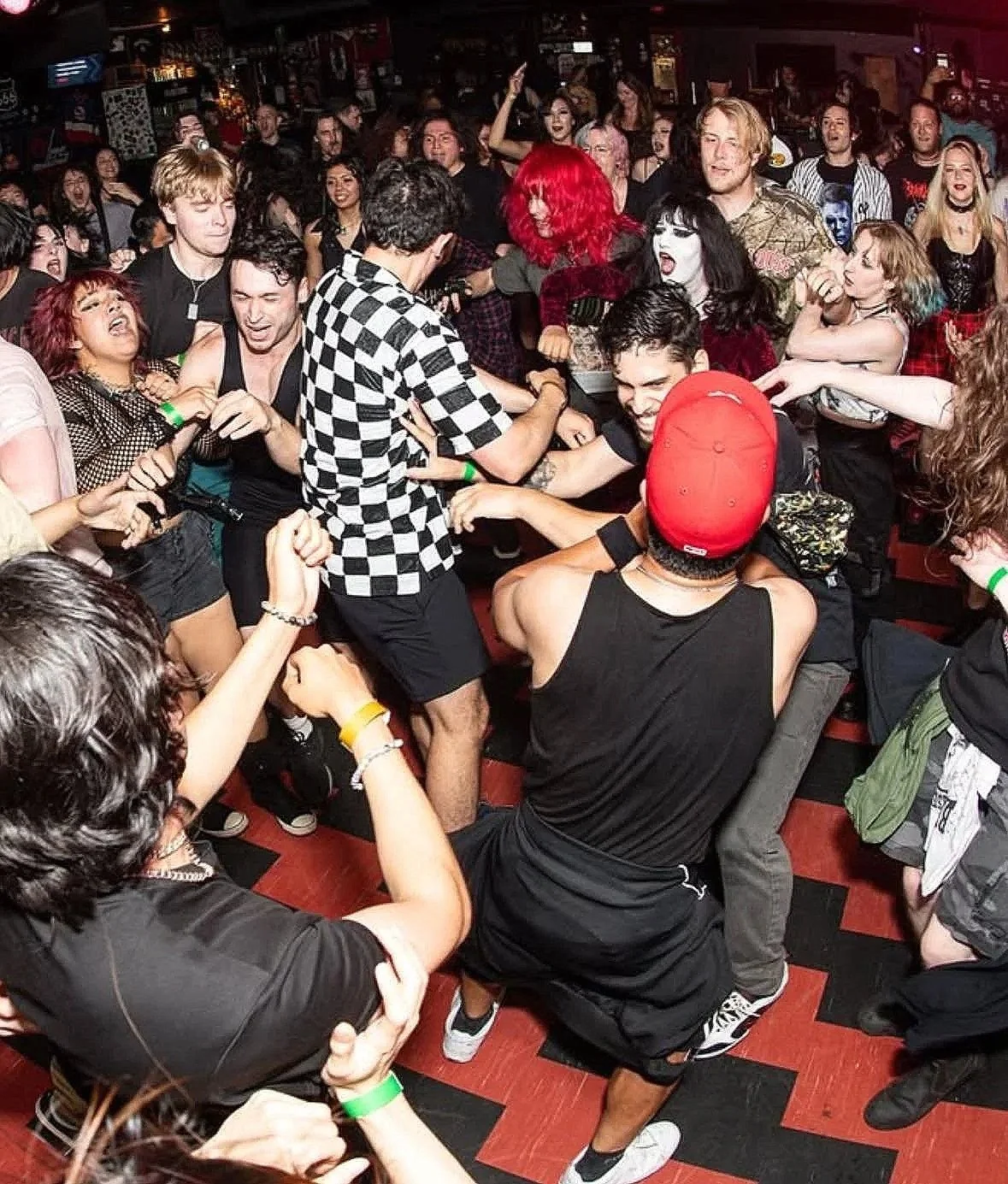A lively scene at a concert or dance event with a large crowd of people dancing and fighting, surrounded by a dark indoor setting with red and black flooring.