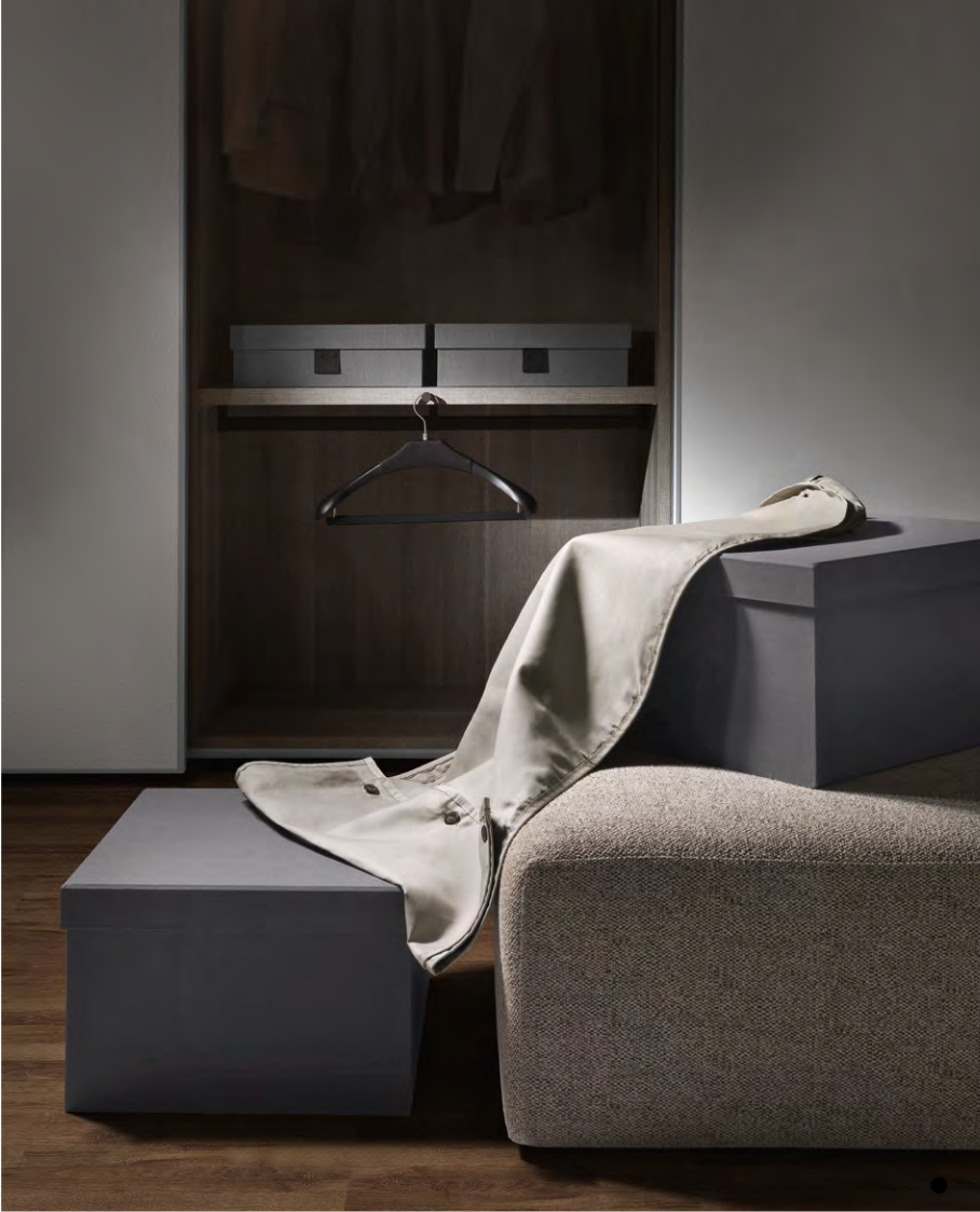 Une pièce avec un placard ouvert, une chaise ou un banc gris, une veste en satin beige déposée dessus, une boîte ou un coffret gris, et un sol en bois.