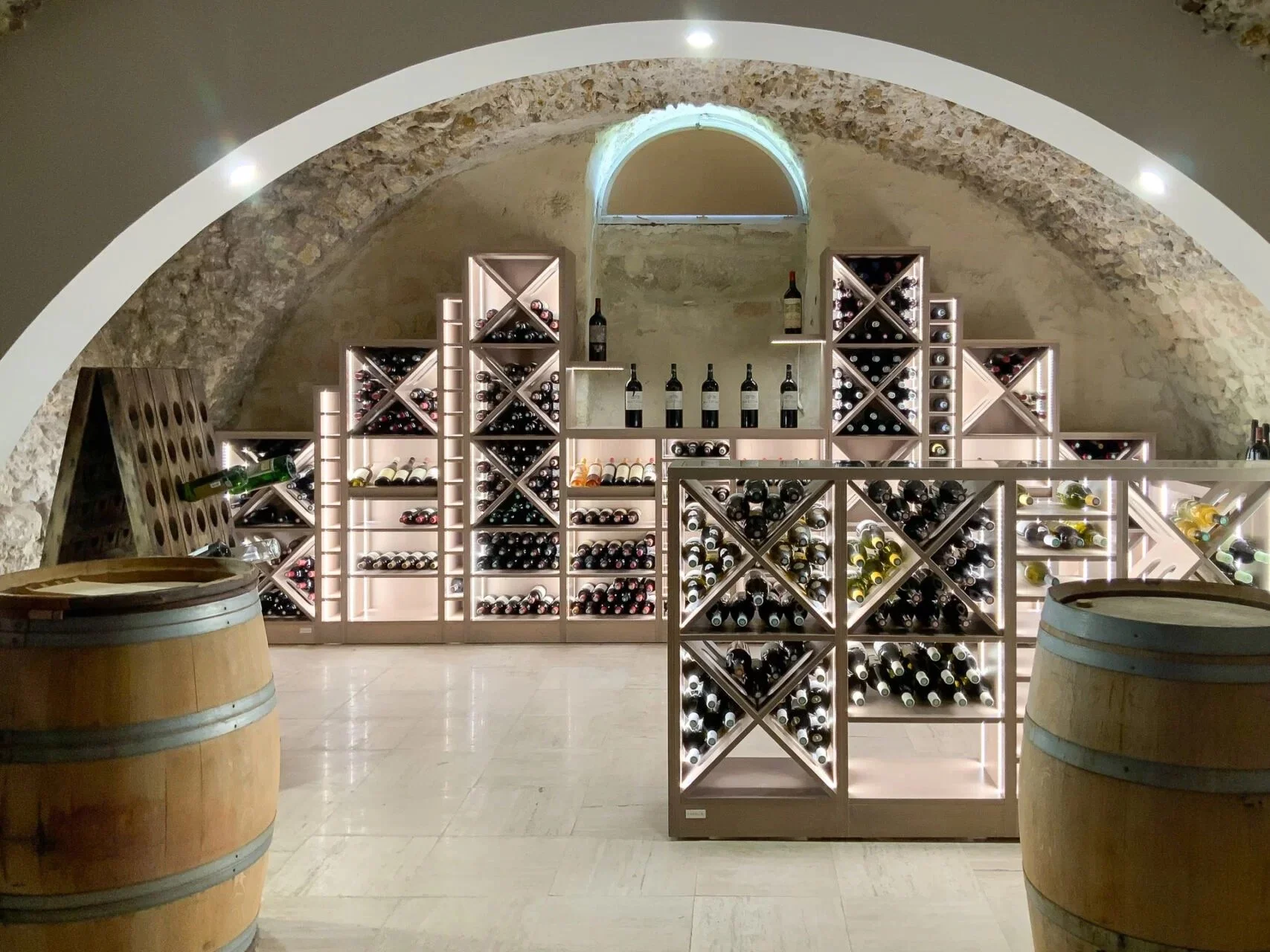 Agencement d'une Cave à vin sur mesure  avec étagères en bois éclairées, remplie de bouteilles de vin, deux tonneaux en bois à l'avant, dans un espace avec un plafond voûté en pierre.