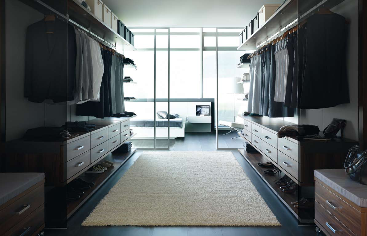 Un dressing Logos Raised avec des vêtements suspendus, des chaussures et des meubles de rangement, avec une chambre en arrière-plan, des portes coulissantes vitrées de séparation et une grande fenêtre apportant de la lumière naturelle.