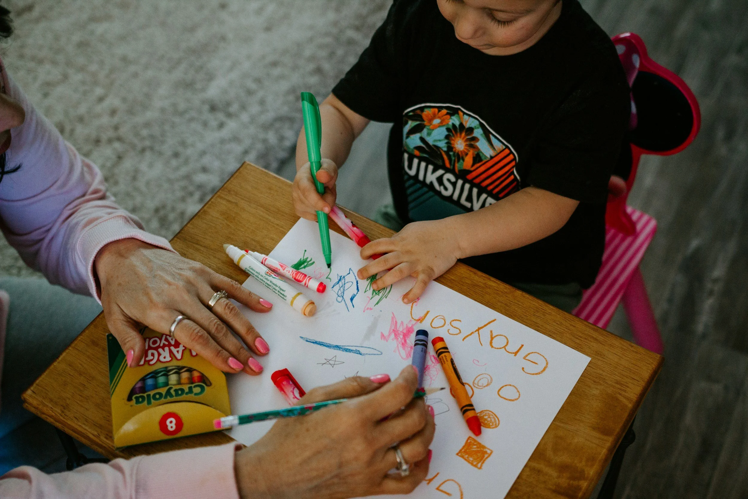 A child coloring on a piece of paper with crayons, surrounded by an adult’s hands and a box of Crayola crayons on a wooden table.